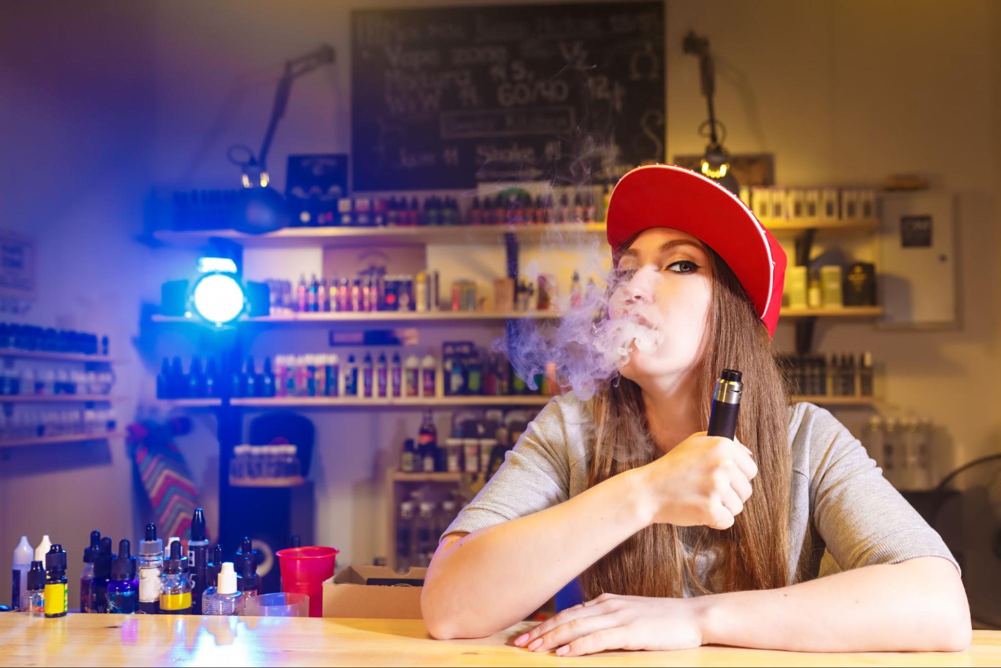 Young, pretty woman in a red cap vapes, with vape storage behind her.
