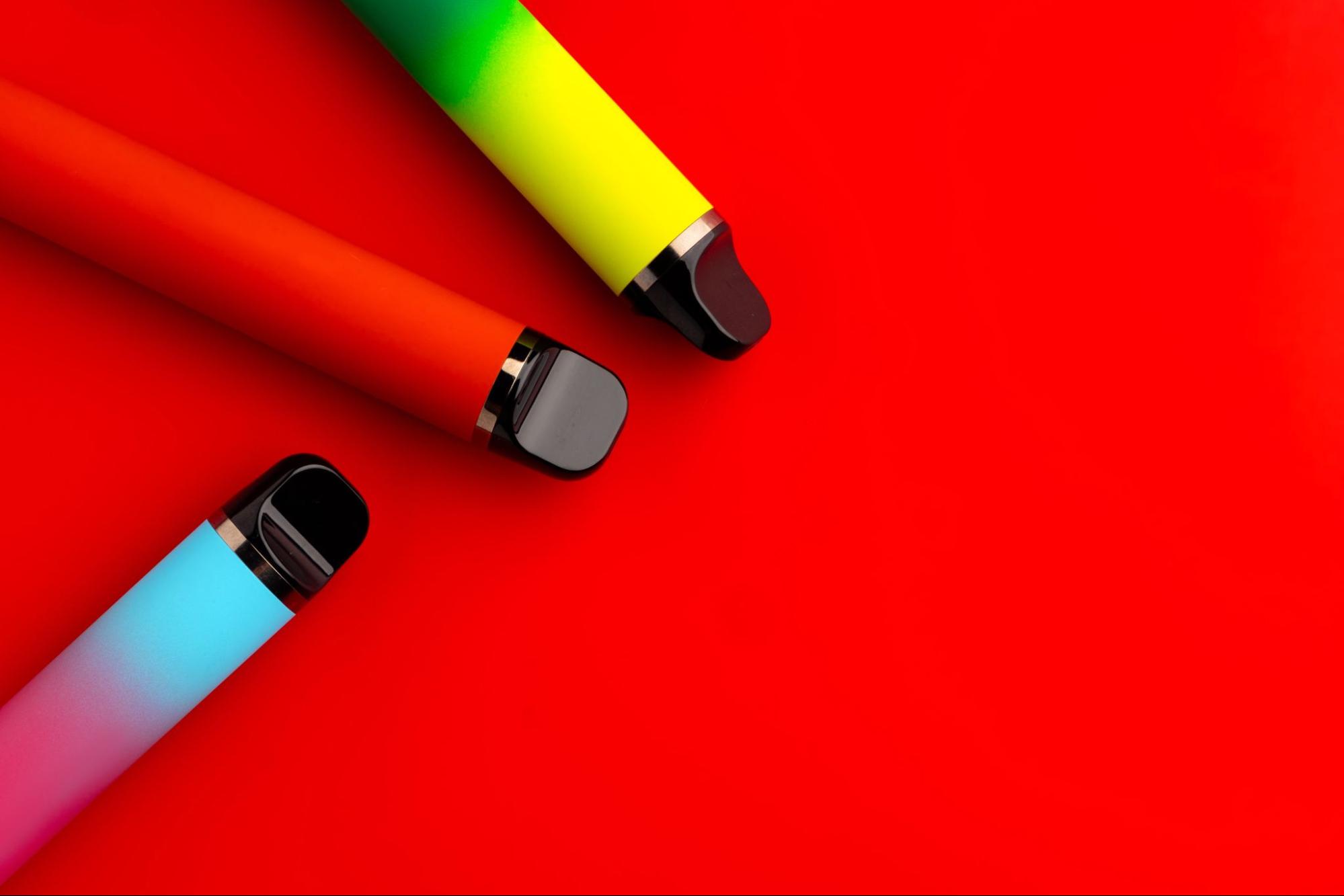 Close-up of disposable vapes in light blue, red, yellow, and green colors against a vibrant red background