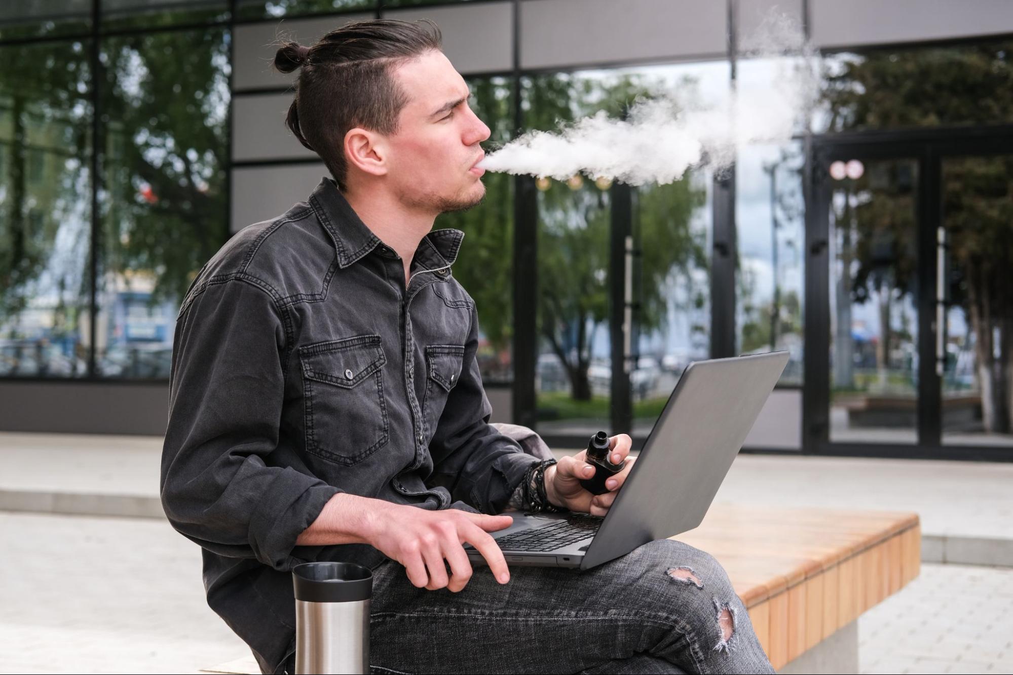 A ponytailed man seated on a bench near the college entrance works on his laptop with a water canister beside him while enjoying a vape.