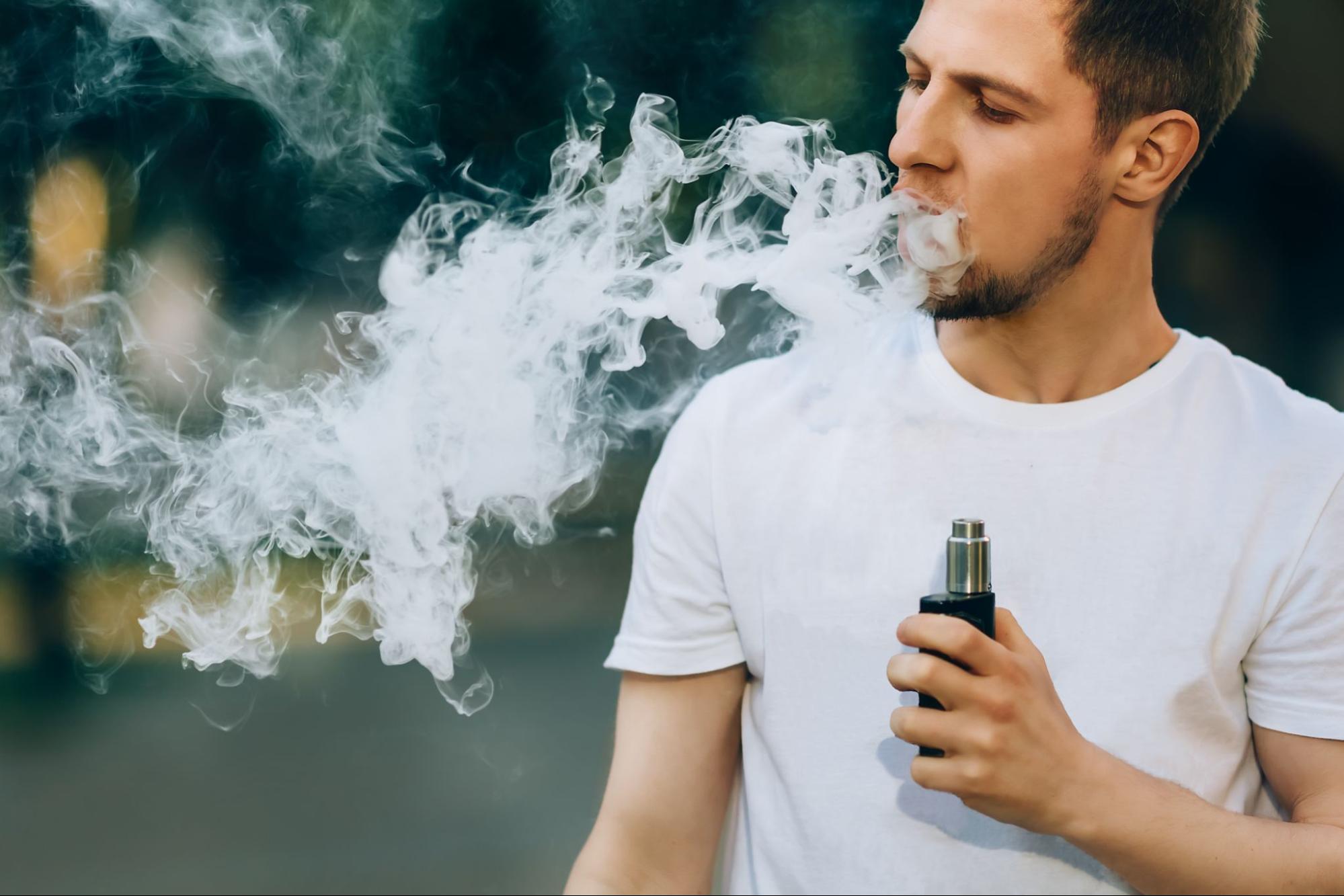 A young bearded man outdoors creates large vapor clouds with his box mod, showcasing the power of vaping technology.