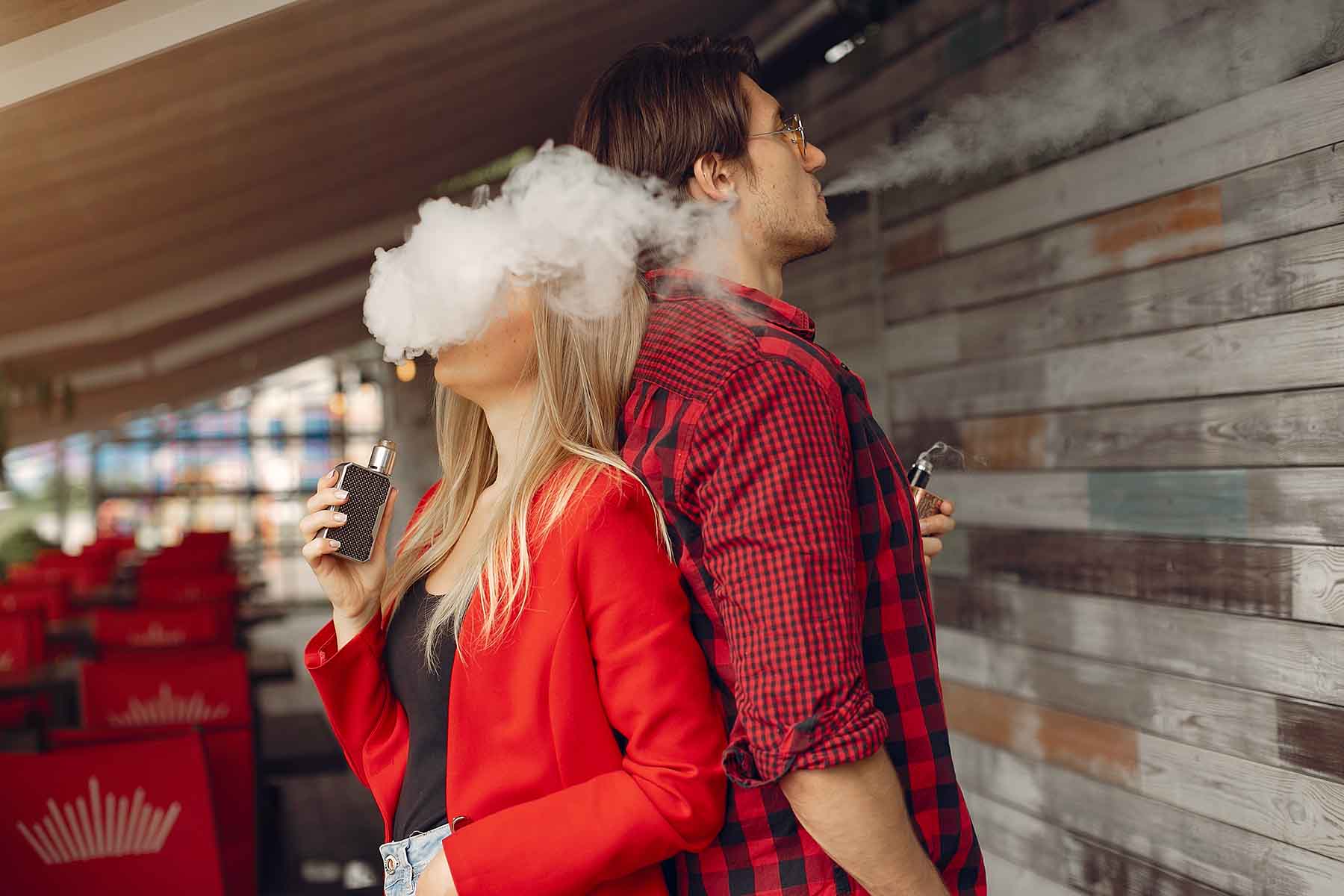 A couple both wearing red standing back to back while both of them are vaping.