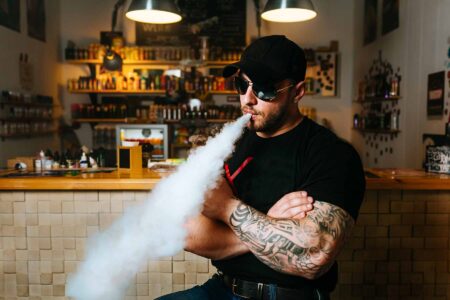A man with tattoos wearing a black hat, black shirt, and sunglasses blowing clouds at a vape store.