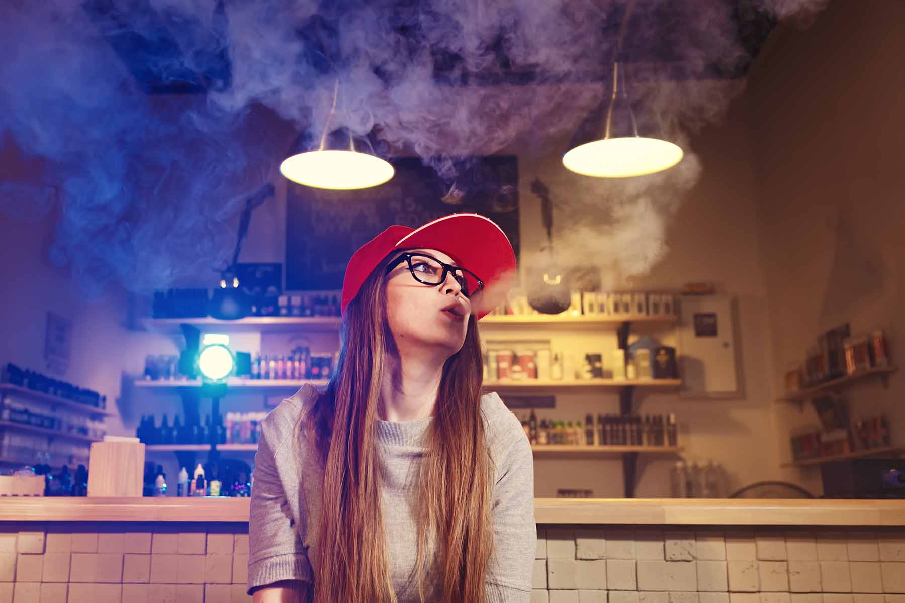 A woman with long brown hair wearing glasses and a red baseball hat blowing out vapor at a vape store.