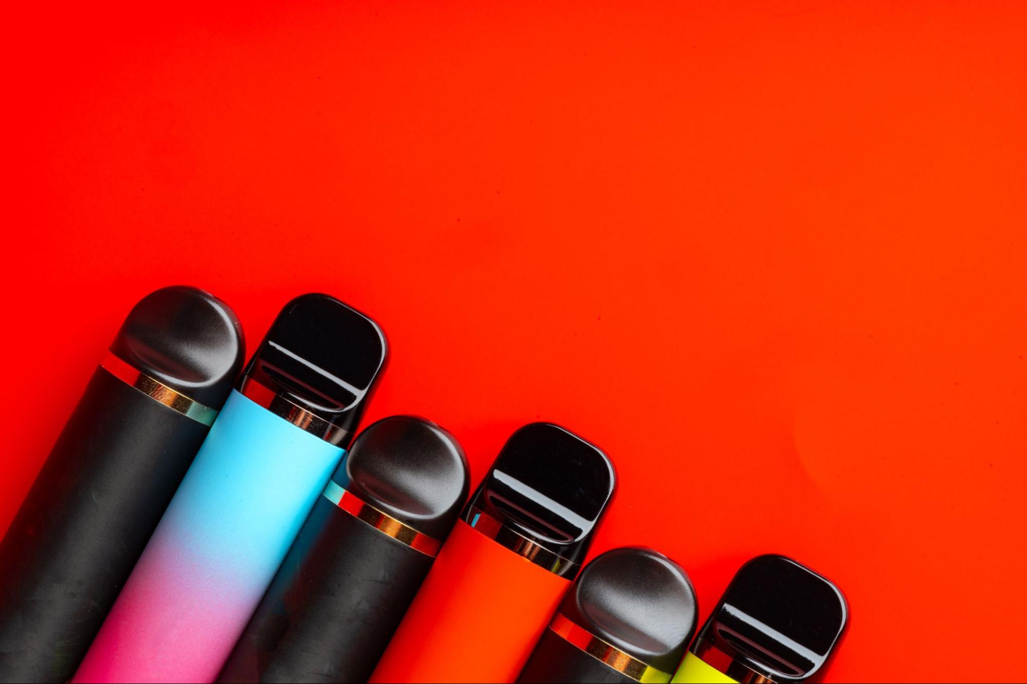 Several different types of disposable vaporizers of assorted colors in a row against a red background.