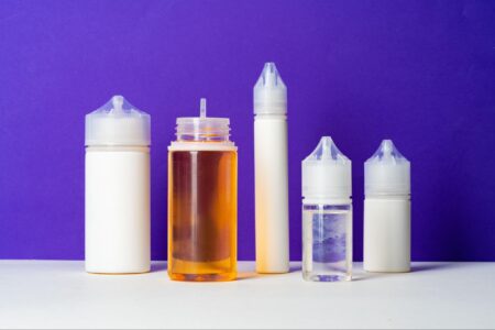 Three white and two transparent e-juice bottles in varying sizes are displayed on a purple background, illustrating the diversity of e-juice sizes.