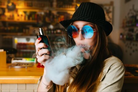 A woman wearing a fedora and oversized sunglasses vaping with a box mod inside of a vape store.