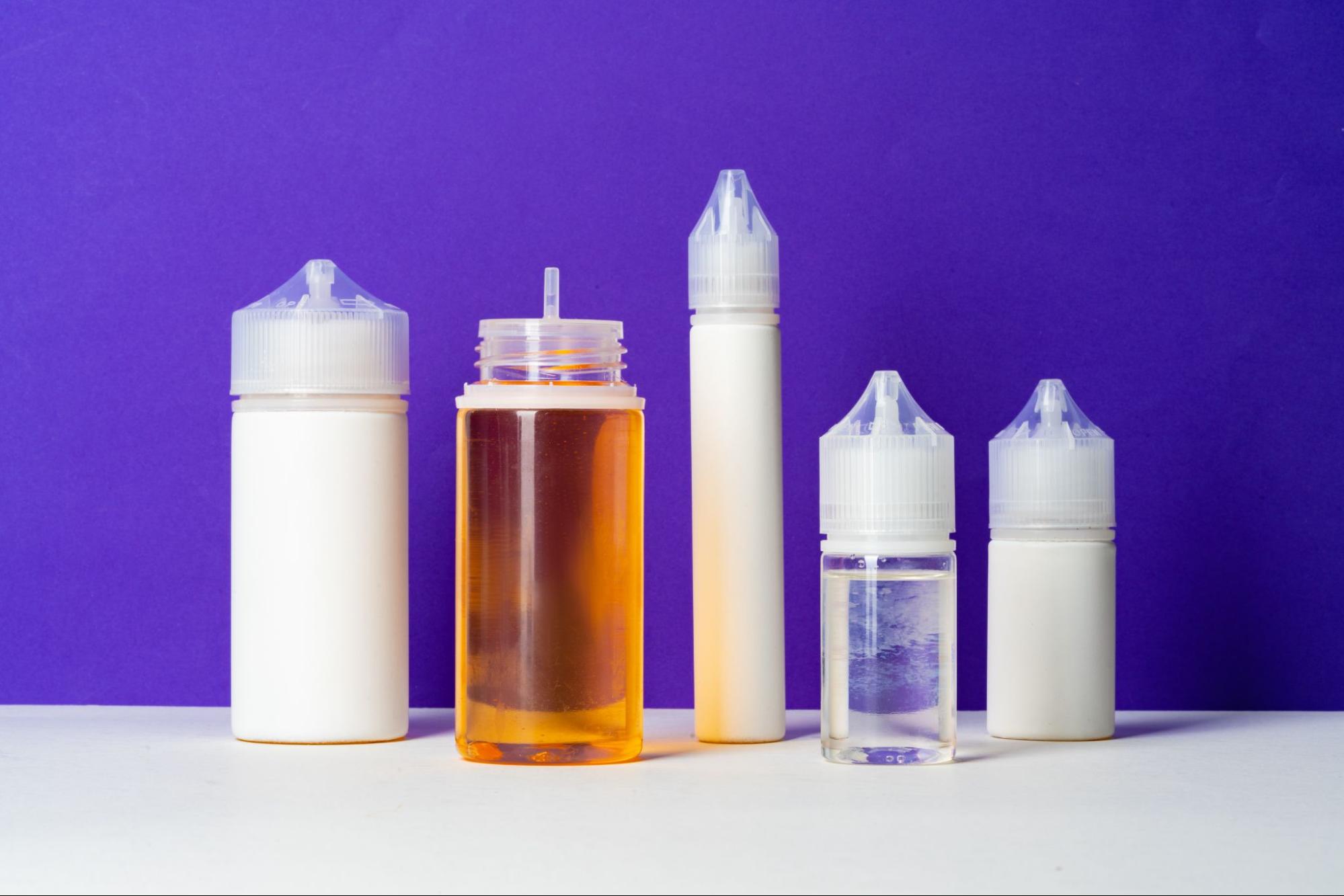 Three white and two transparent e-juice bottles in varying sizes are displayed on a purple background, illustrating the diversity of e-juice sizes.