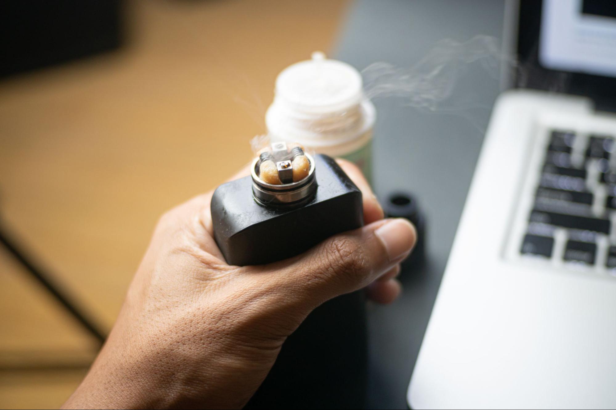 A cropped photo of a man holding down the button for the battery on a box mod vape with a buildable atomizer.