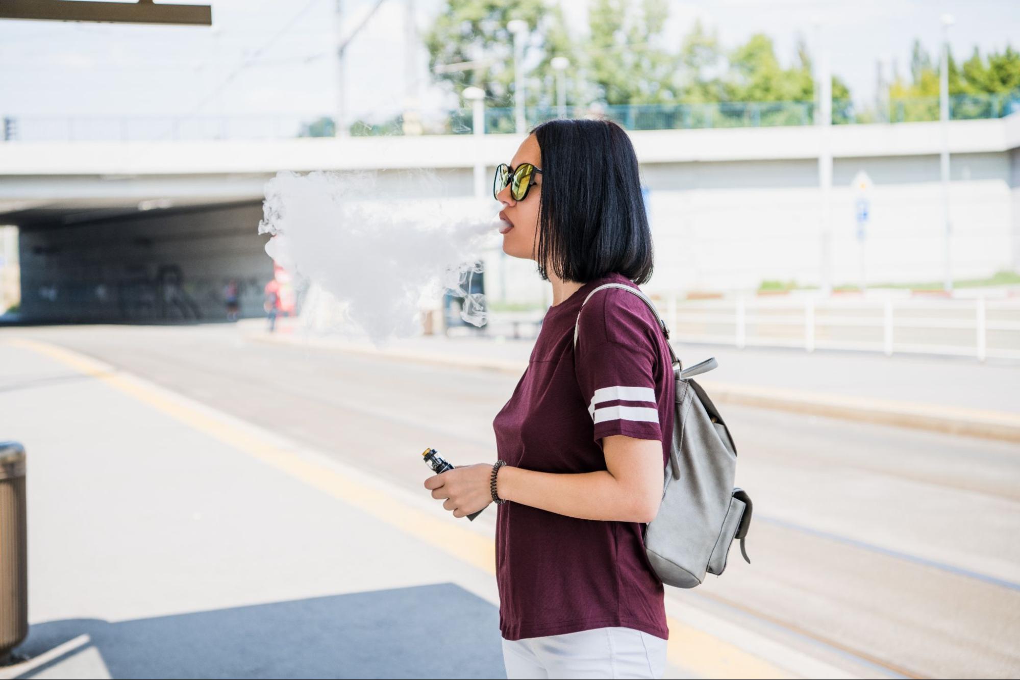 A young woman vapes an e-cigarette outdoors. 