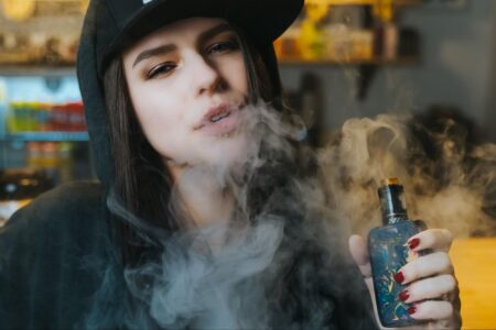 A close-up view of a woman wearing a black hat and hoodie holding a vape mod and blowing vapor towards the camera.