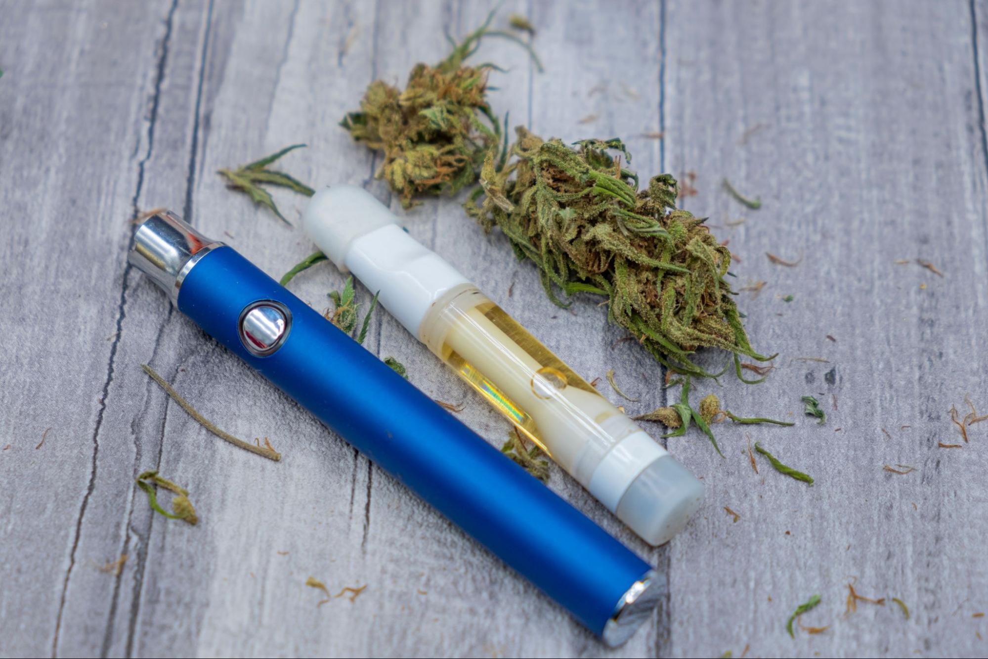 A cropped view of a blue vaporizer pen next to a cartridge and ground marajuana on top of a wooden surface.