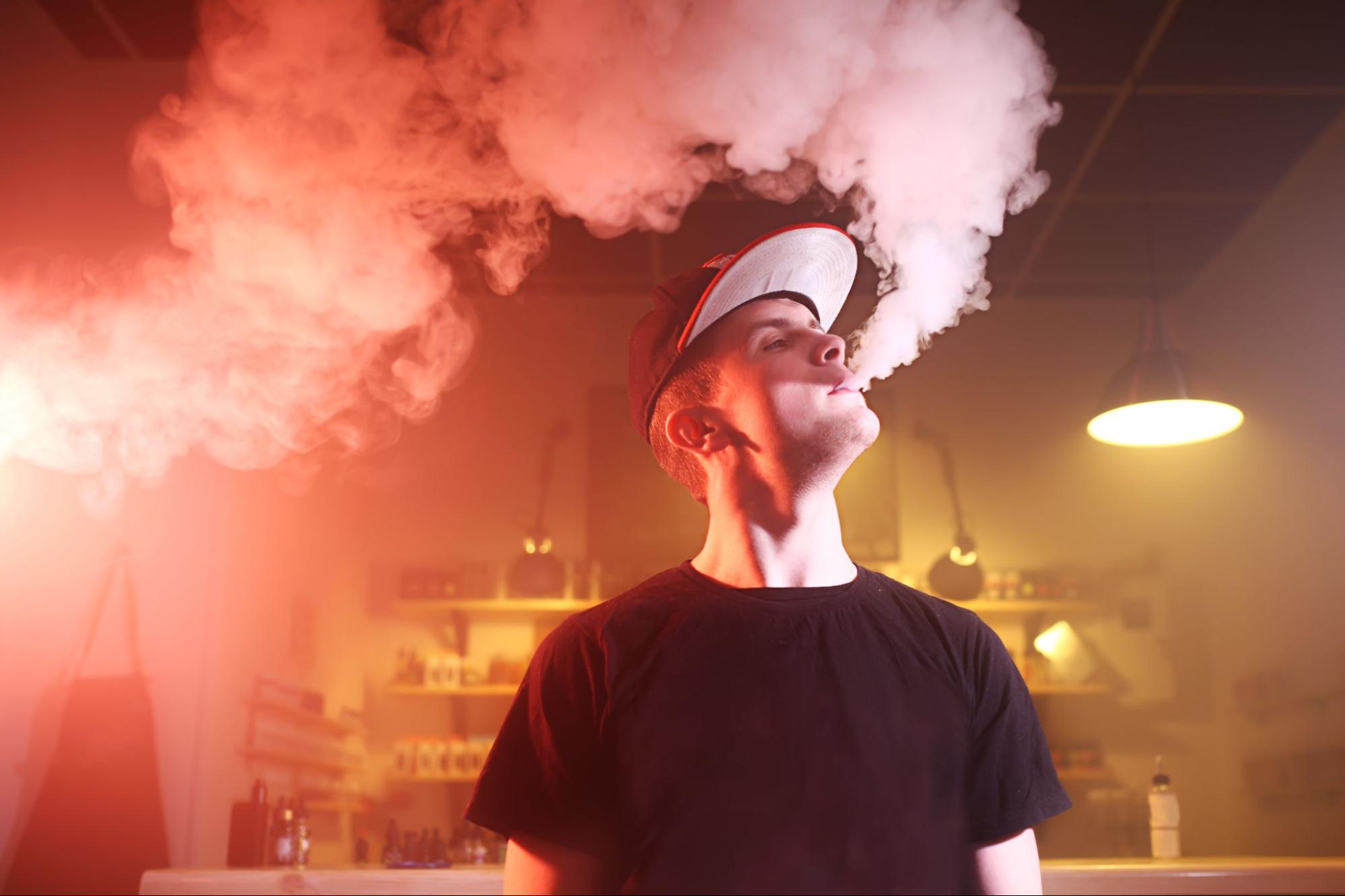 A man in a baseball hat and black t-shirt exhales a big vapor cloud under red and orange lights inside a vape store.