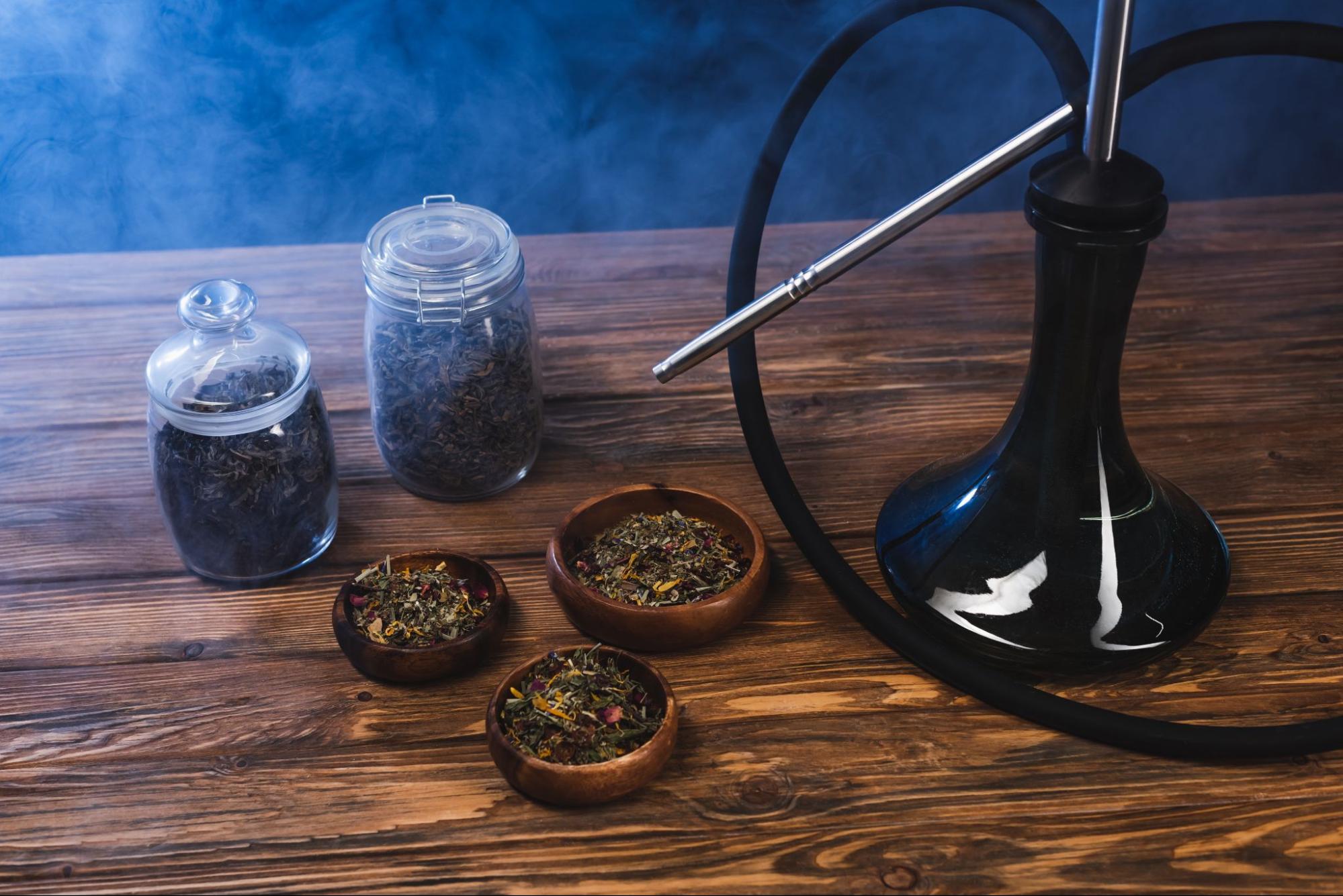 Dry tea in jars and bowls sits near a hookah on a wooden surface against a black background, surrounded by smoke.