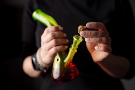 Cropped view of a woman’s hands as she puts marijuana into a bong bowl against a dark background.