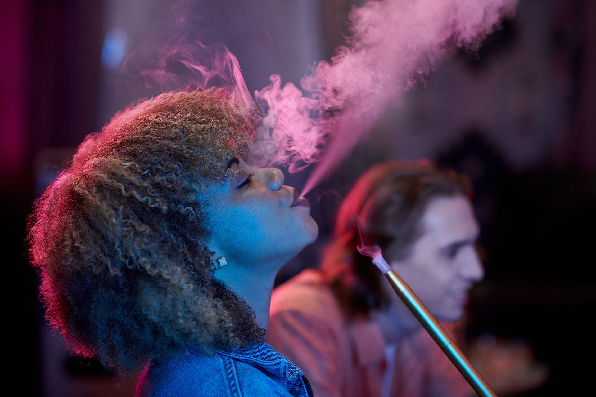 A woman sitting next to a man with long hair blowing smoke out of her mouth after taking a hit of her vape device.