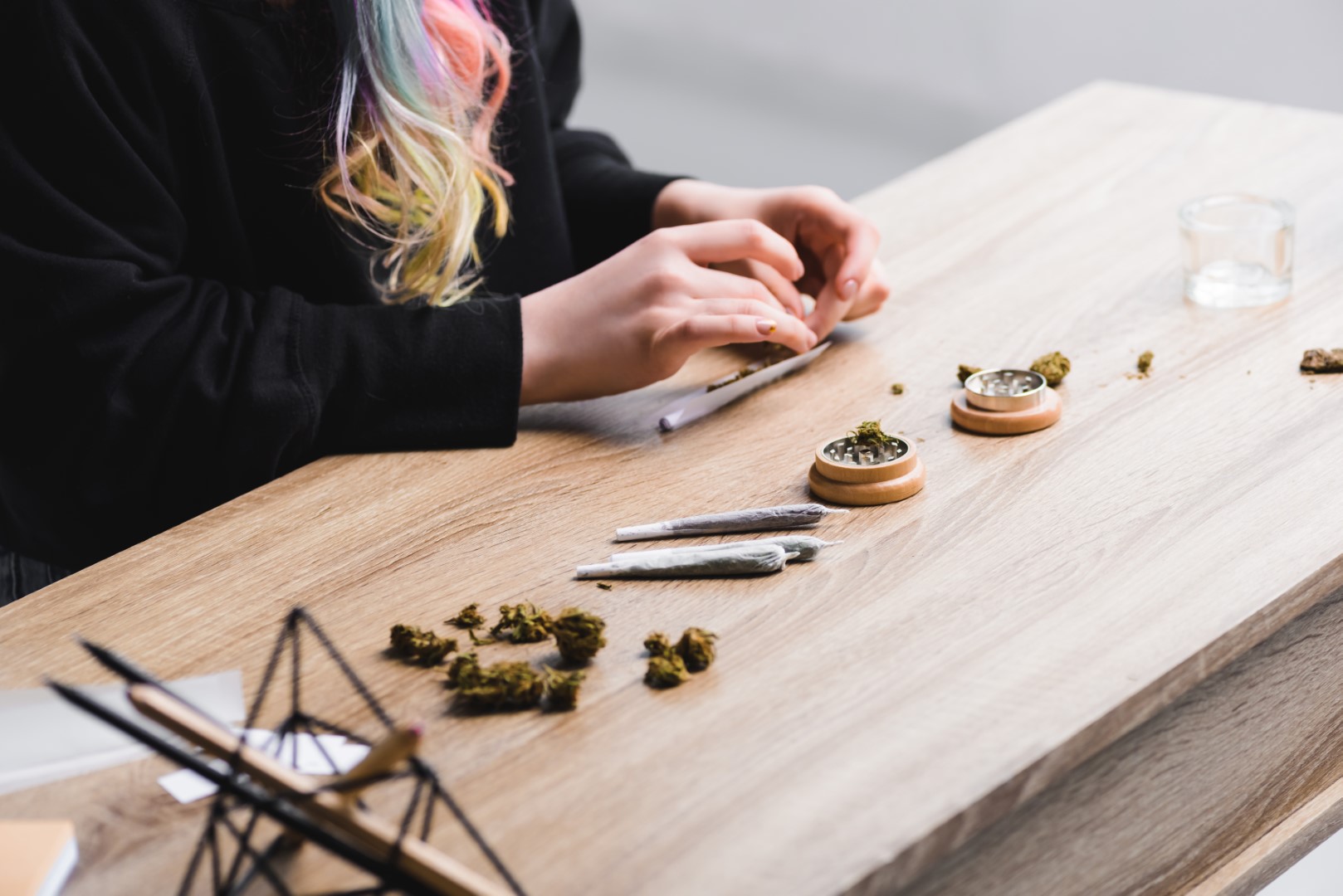 A cropped view of a woman rolling a joint on a table next to whole buds, joints, a grinder, and a glass jar.