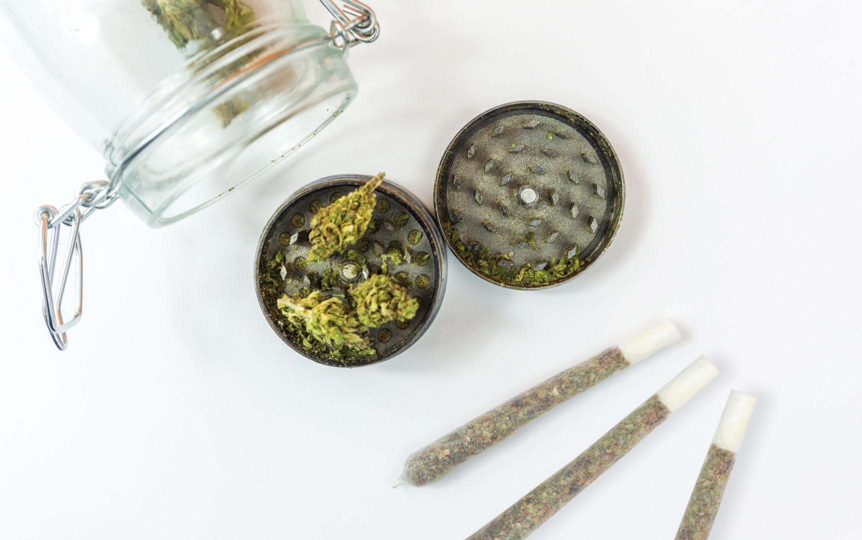 A glass jar with cannabis inside, three rolled joints, and a back grinder with one-half of whole buds on top and the other half with some ground weed on top
