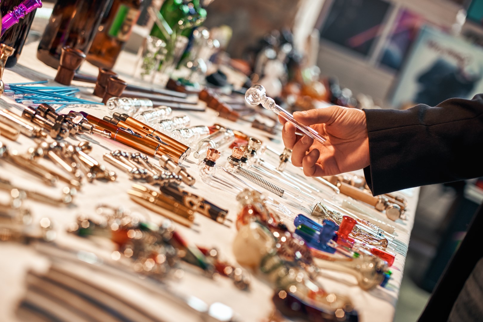A wooden table with many types of marijuana smoking devices and a person’s hand holding one of the glass pipes.