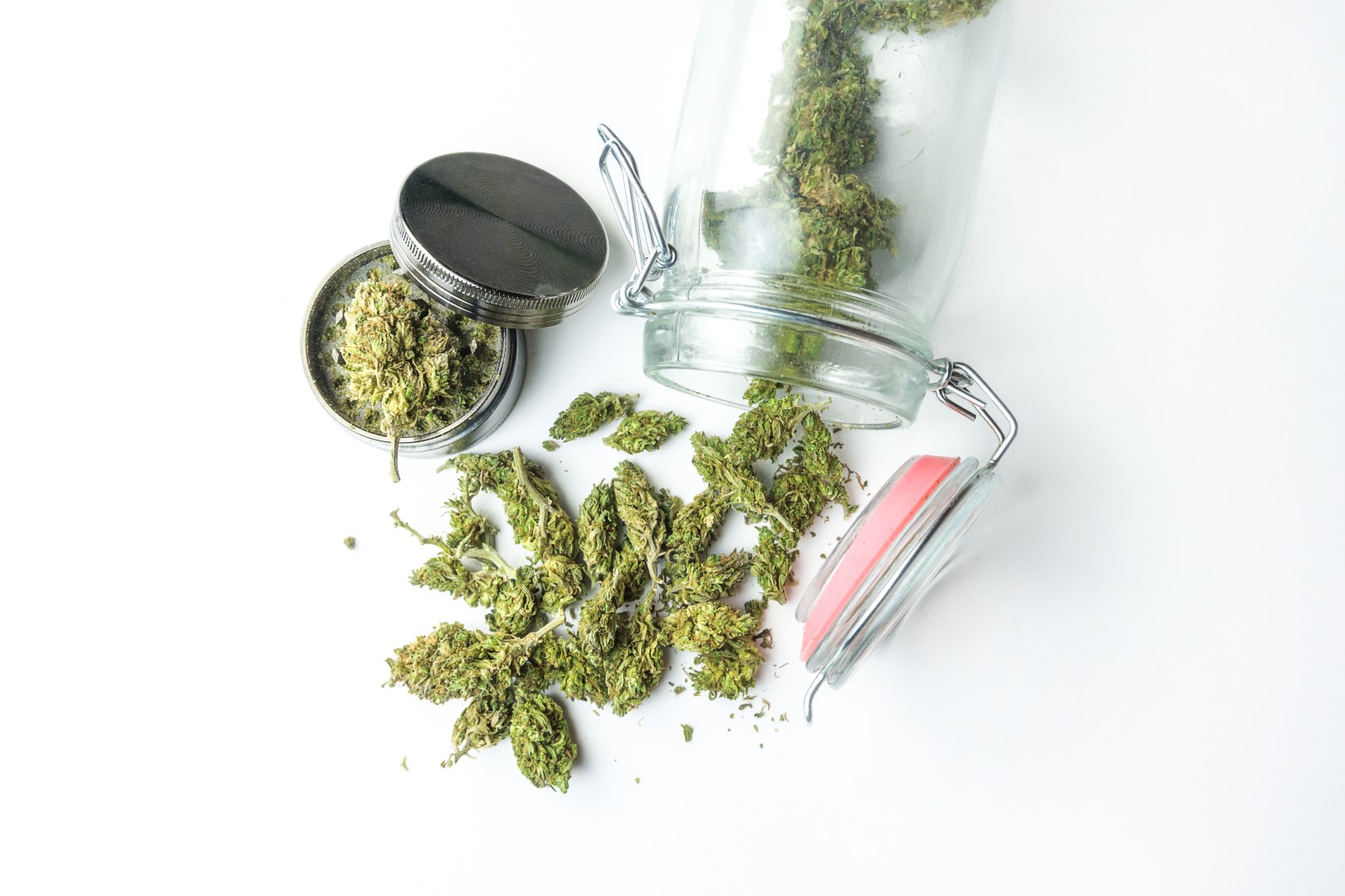 An open weed grinder with ground buds inside next to a glass jar of weed tipped over with whole buds falling out against a white background.