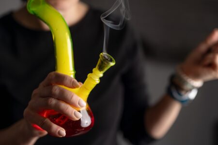 Cropped photo of a woman’s hand holding a yellow and red bong in one hand against a blurry background.