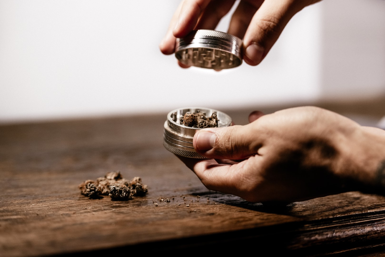 Cropped view of a person’s hands using a grinder with ground weed inside and whole buds on the wood table.