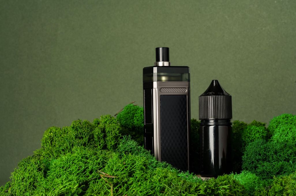 Vape device next to liquid on a green plant.