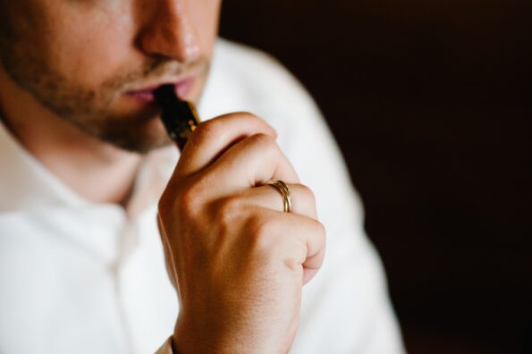 A close-up cropped view of a man in a white shirt holding a vaporizer up to his mouth against a black background.