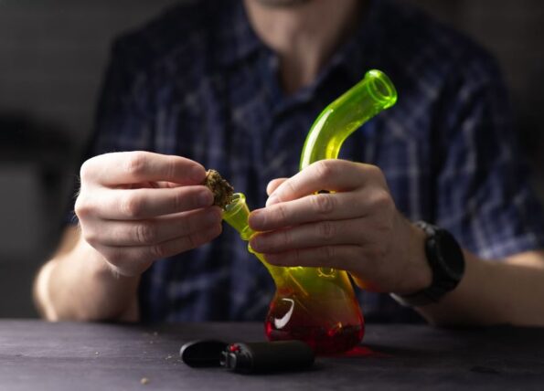 A cropped view of a man’s hands holding a red, yellow, and green miniature bong while packing the bowl with cannabis bud.