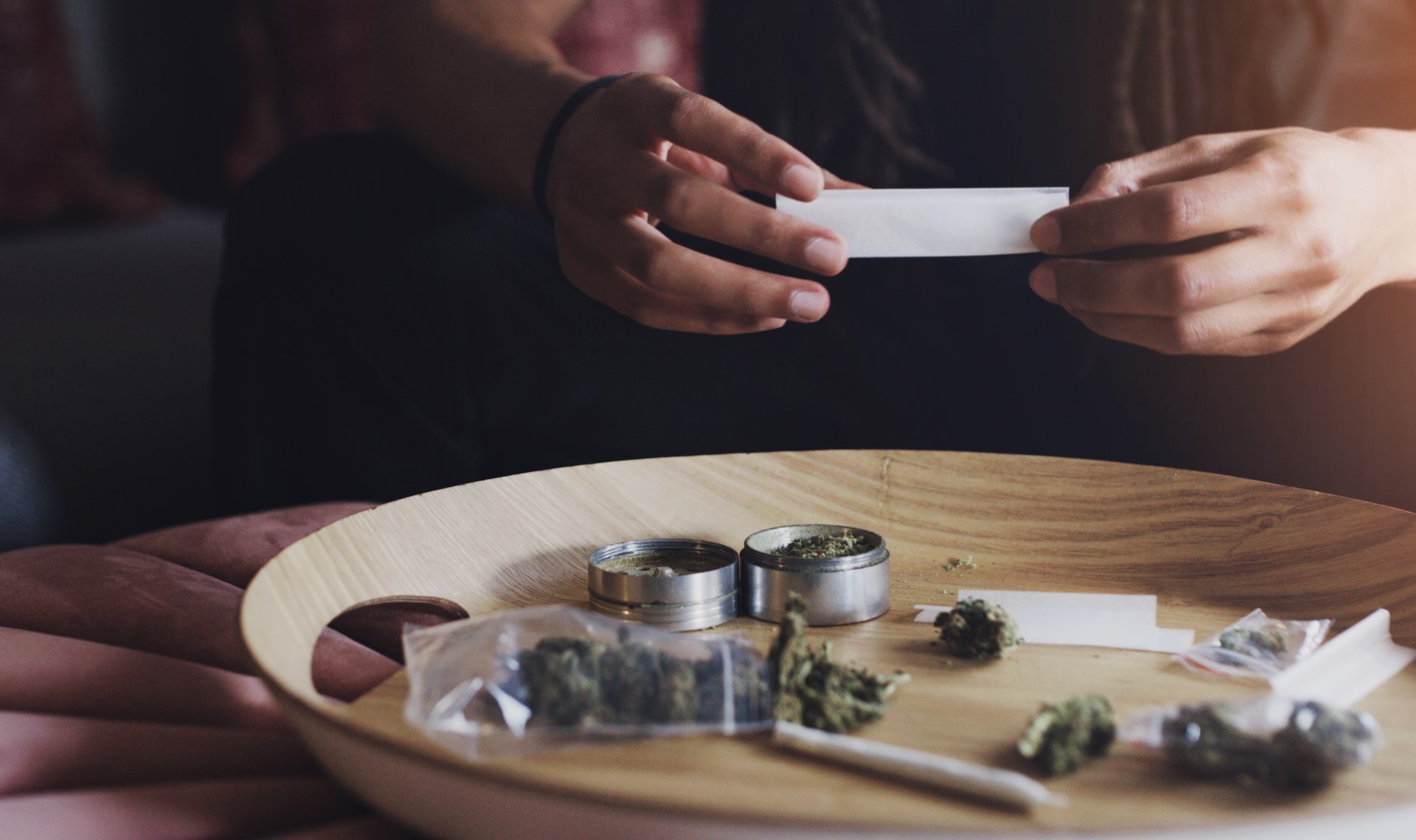 A cropped view of someone’s hands rolling a joint over a wooden rolling tray with a grinder, marijuana, and papers on top of it.