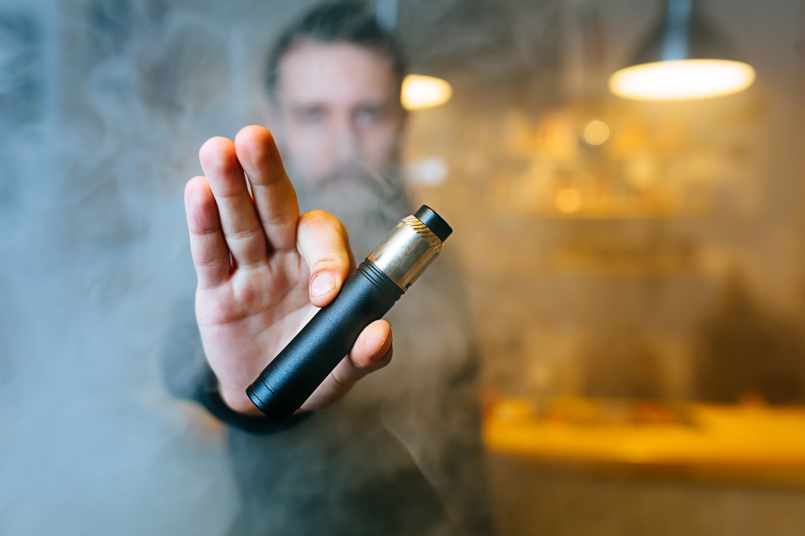 A man with a beard holding a pod system vape out to the camera with him obscured by vapor smoke in the background.