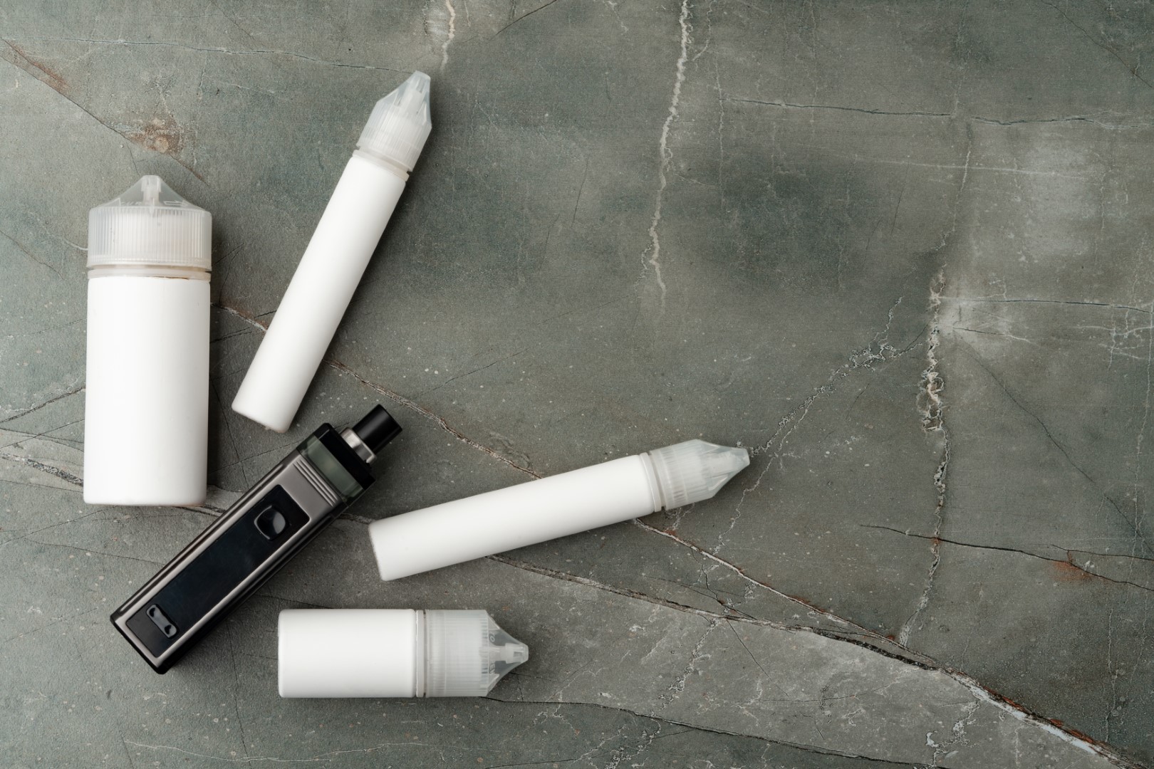A vaporizer and four white bottles of vape juice of different sizes against a gray stone background.