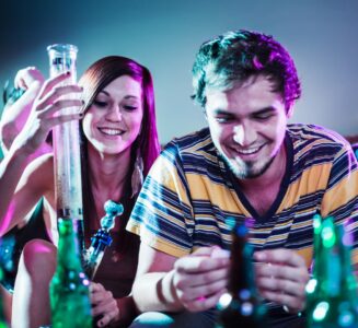 A woman and a man smile at a party while she holds a large bong, and he rolls a joint.