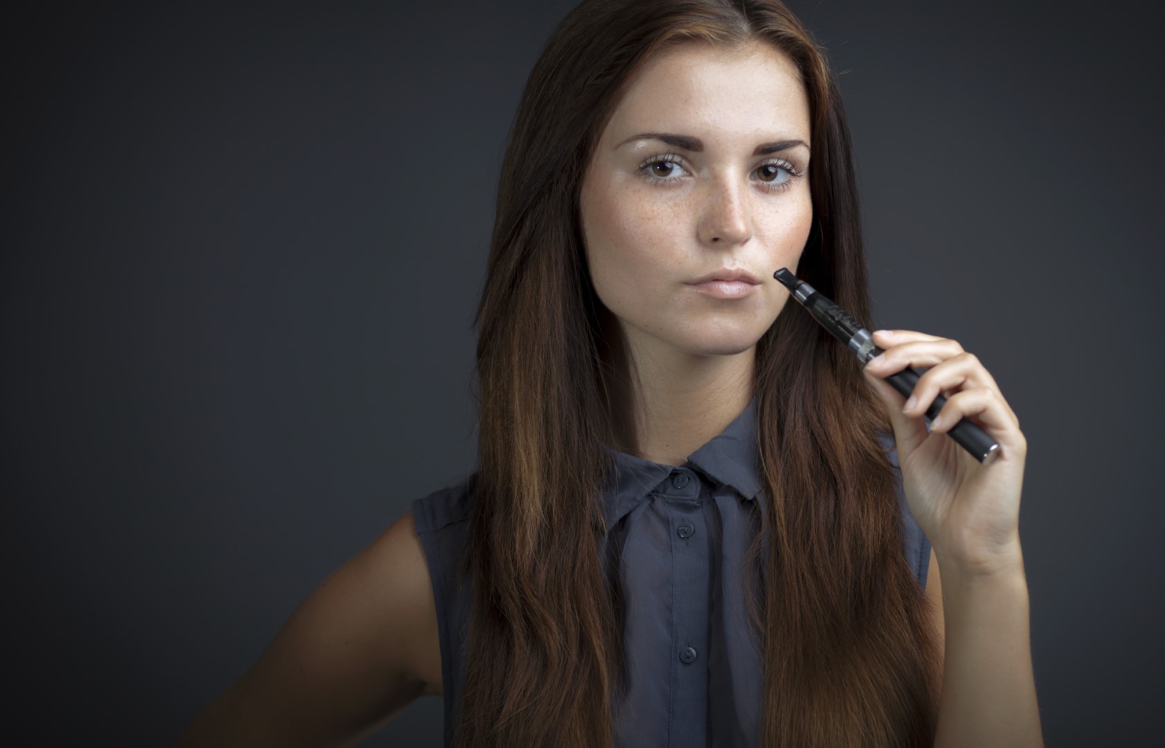 A woman with long brown hair is wearing a gray top and holding a vaporizer up to her mouth.