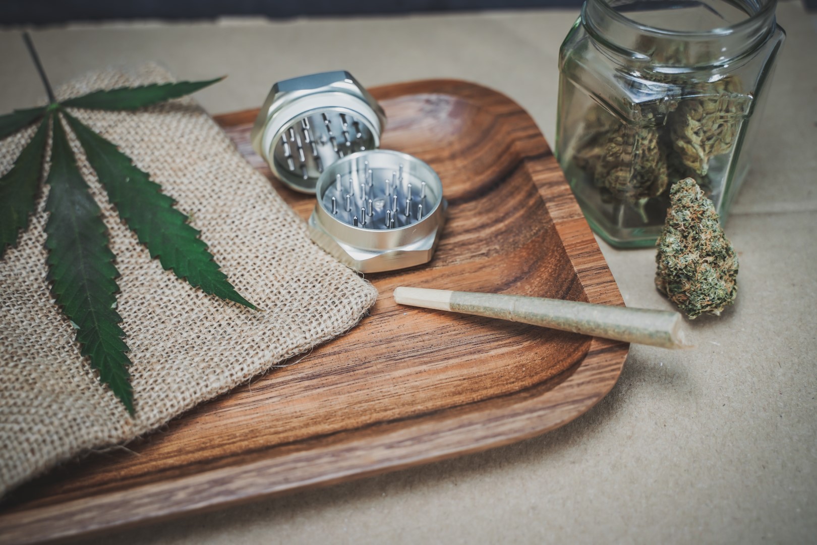 A wooden rolling tray with a hemp cloth, a grinder, and a joint on top of it next to a glass jar of marijuana.