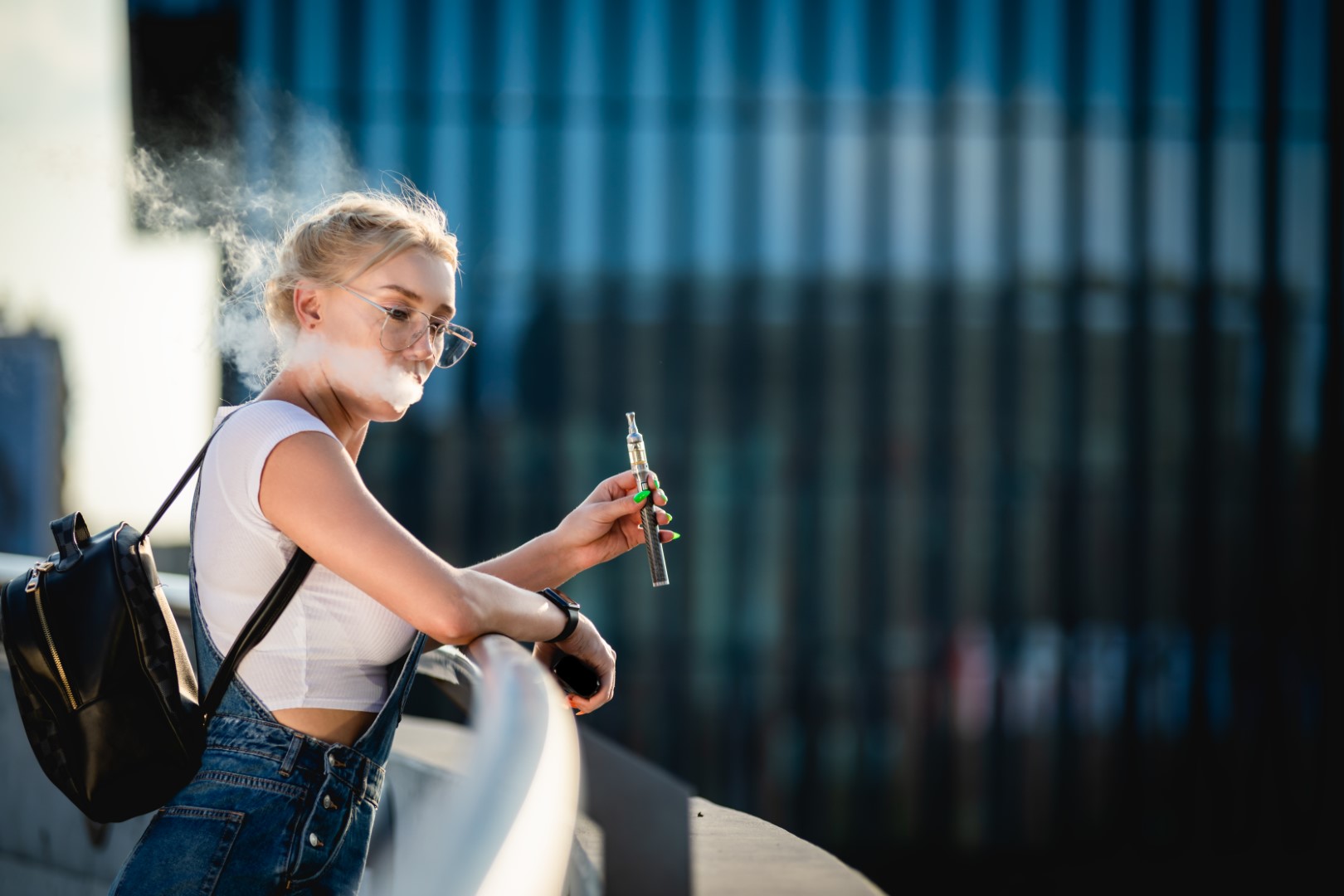 A young woman with blonde hair and glasses wearing a white crop top, overalls, and a black backpack leaning over a balcony while holding a vaporizer and blowing out vapor smoke.