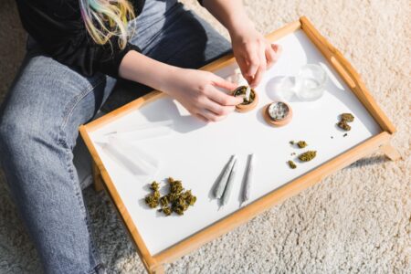 View from above of a woman using a rolling tray, rolling papers, and a grinder to roll joints on the floor.