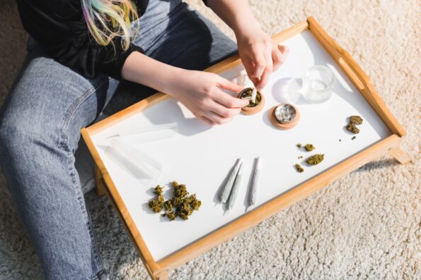 View from above of a woman using a rolling tray, rolling papers, and a grinder to roll joints on the floor.