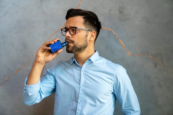 A man with brown hair and glasses is holding a vape with blue vape skin up to his lips.