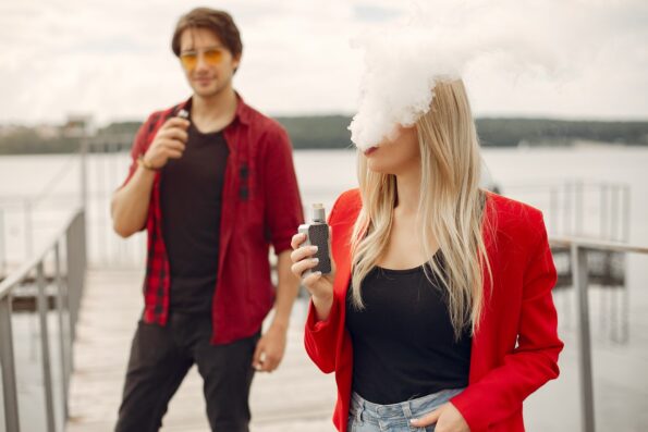 A woman with blonde hair and a man with brown hair, both wearing black shirts and red jackets, vaping with box mods on a dock.