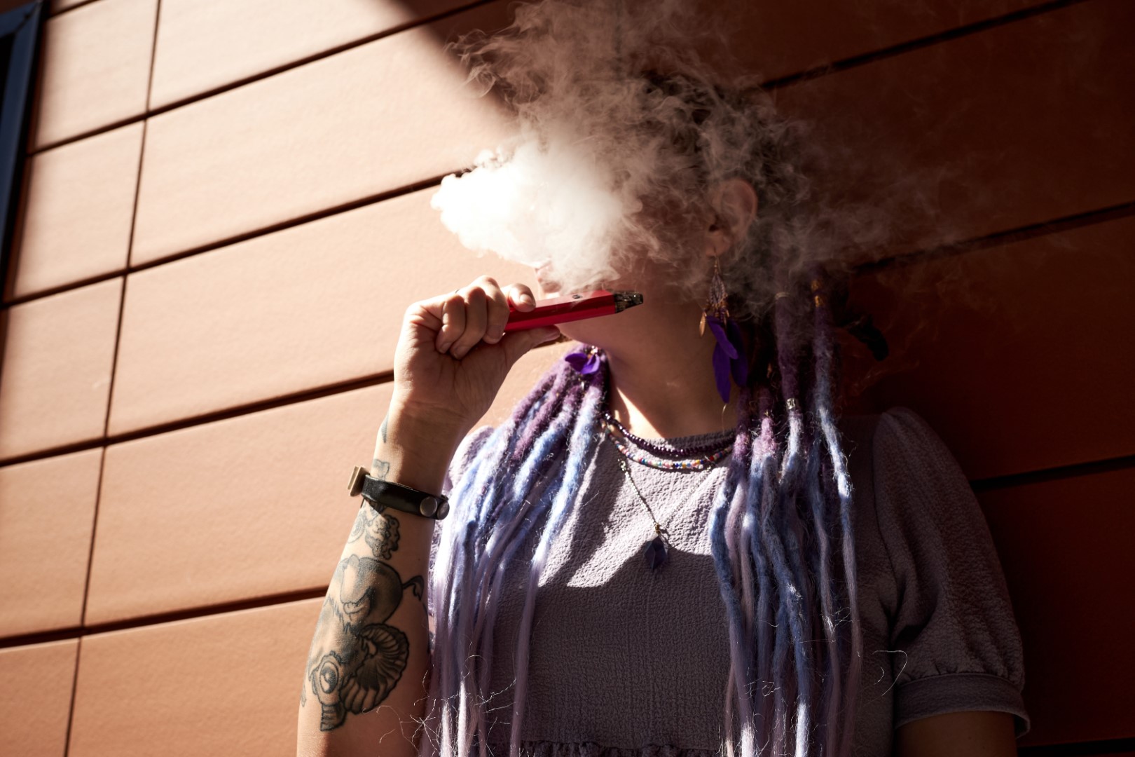 A woman with purple dreadlocks and tattoos vaping with a wax pen, blowing a cloud of smoke out of her mouth with her face obscured. A woman with purple dreadlocks and tattoos vaping with a wax pen, blowing a cloud of smoke out of her mouth with her face obscured.
