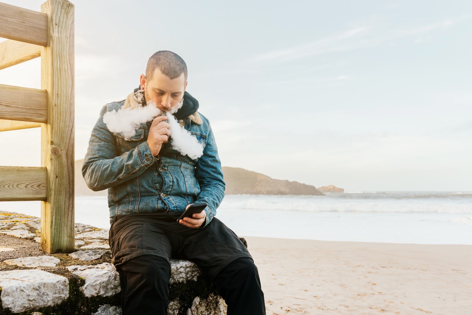 A man with buzzed brown hair wearing a hoodie under a jean jacket holding a vape up to his mouth while blowing out large clouds of vapor while looking down at his phone near a beach.