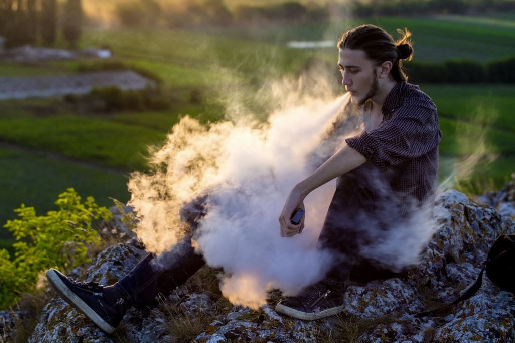 A man sitting on a rock outside smoking from a vaporizer.