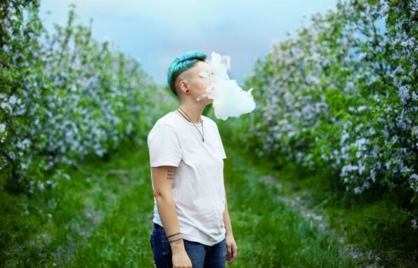 A person exhaling smoke outside beside grass and bushes.