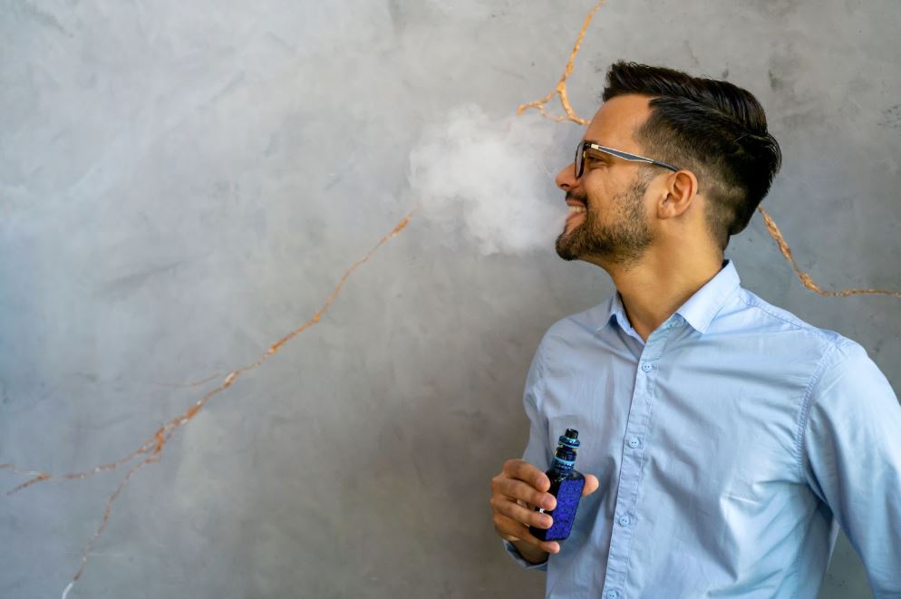 Man in a buttoned shirt releasing smoke out of his mouth while holding a vape device.
