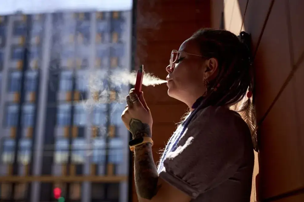 Woman leaning against a wall and holding a vape while exhaling smoke.