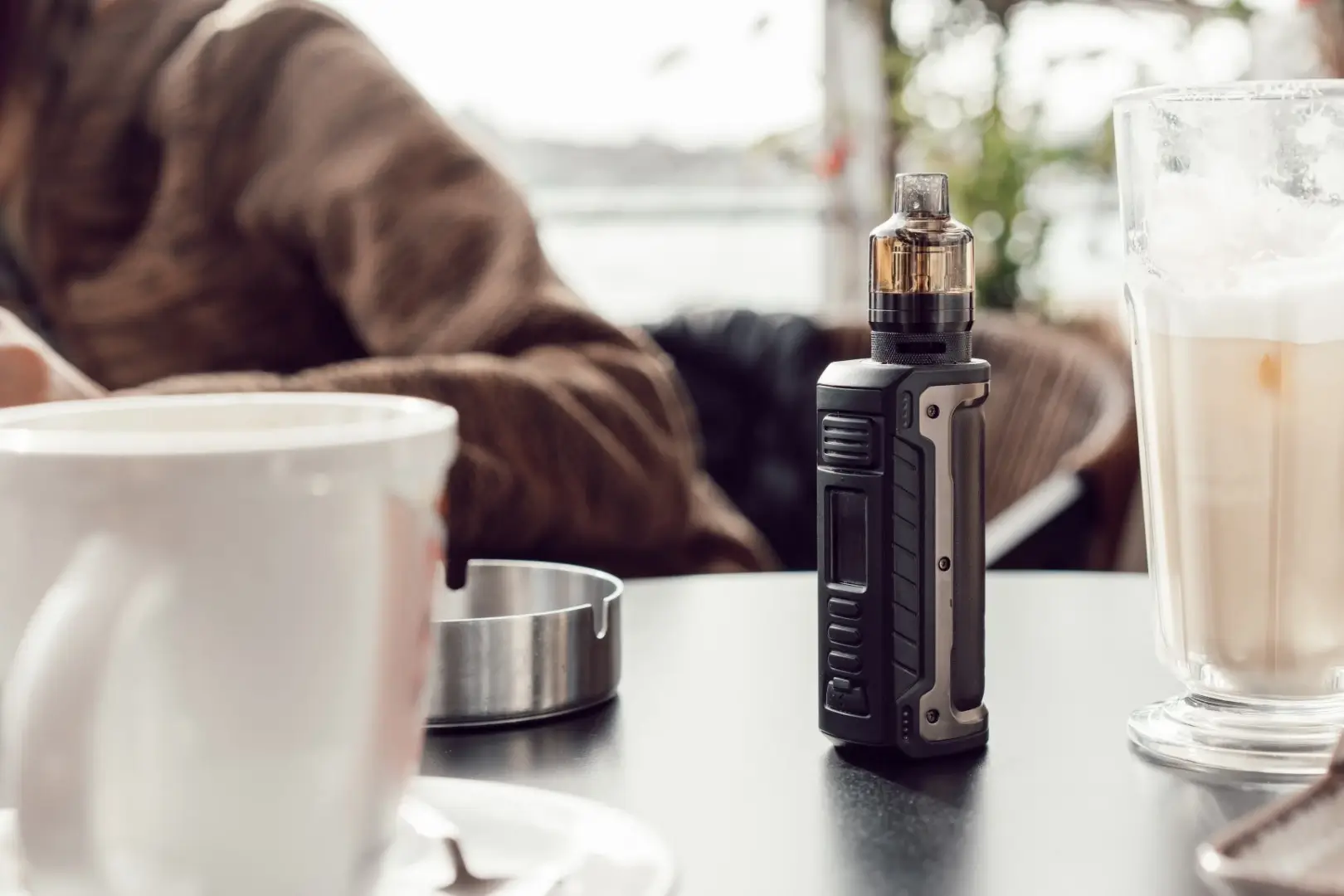 A box mod vape standing on a table at a cafe surrounded by an ashtray and cups with a person leaning out of the frame.