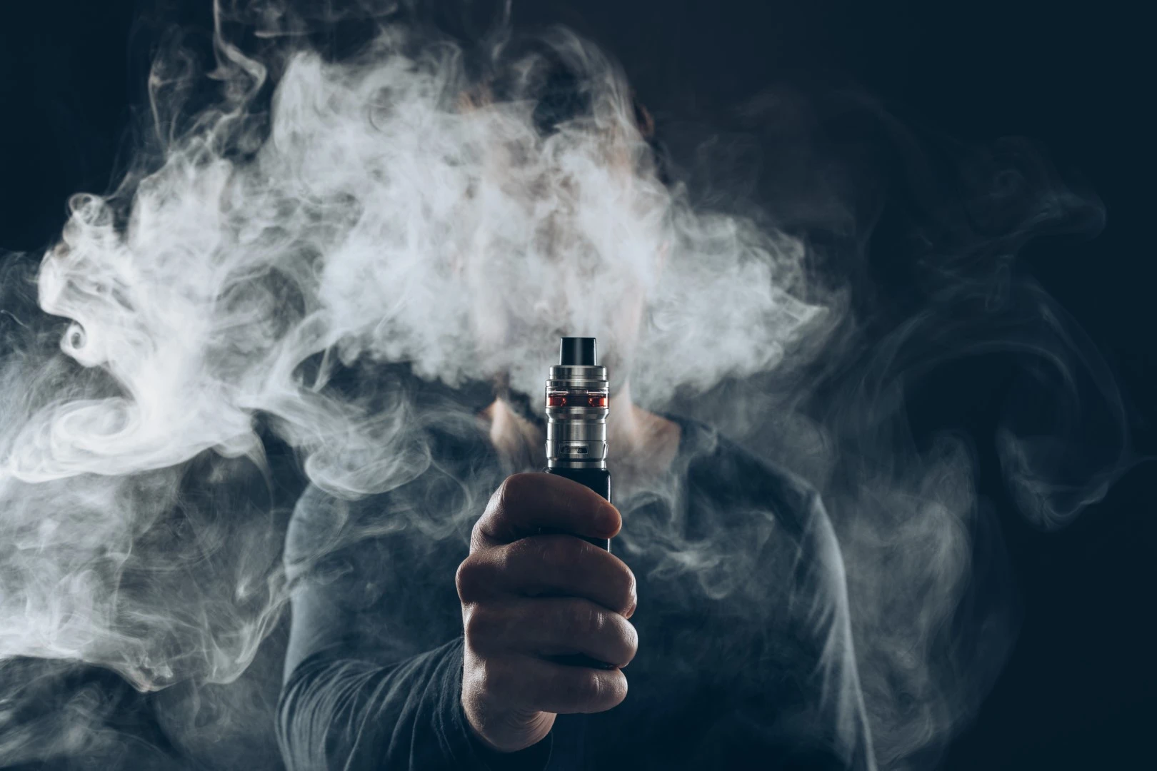  A man holding his vape device in front of the camera while he is obscured by vapor in the background.