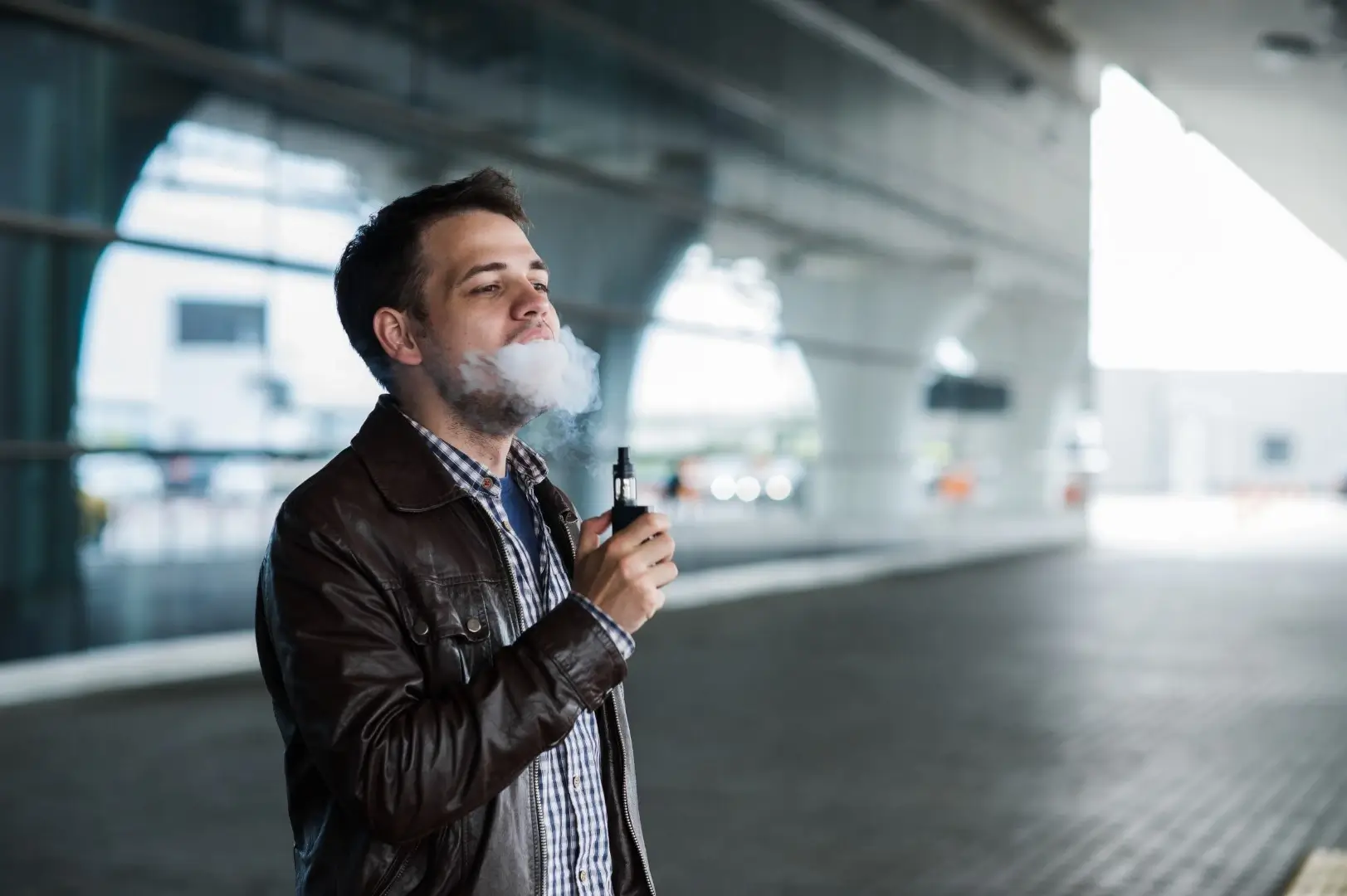 A man wearing a leather jacket and a button-down shirt standing in an airport terminal while vaping.