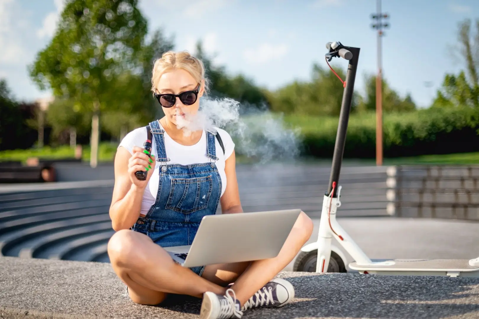 A woman wearing a white t shirt and overalls is sitting on the ground beside a scooter working on her laptop and vaping.