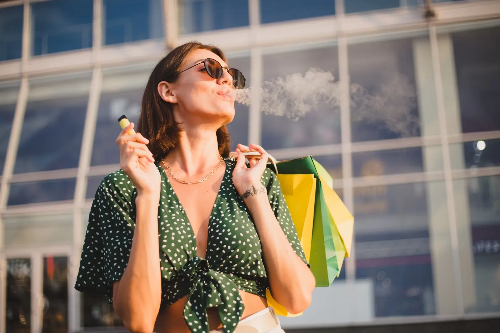 A woman with short brown hair is wearing sunglasses and a green crop top holding shopping bags over her shoulder and vaping.